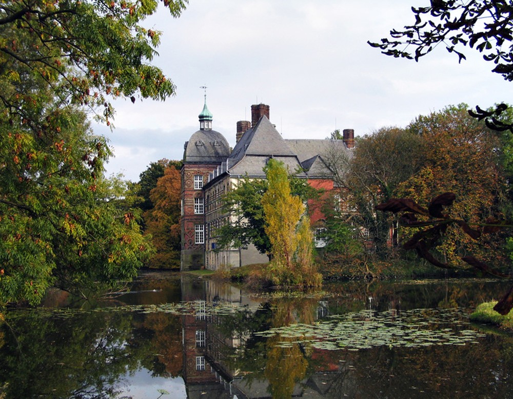 Blick über die Gräfte auf Schloss Hovestadt. Auf dem Wasser befinden sich Seerosen. Am Ufer befinden sich zahlreiche Pflanzen.