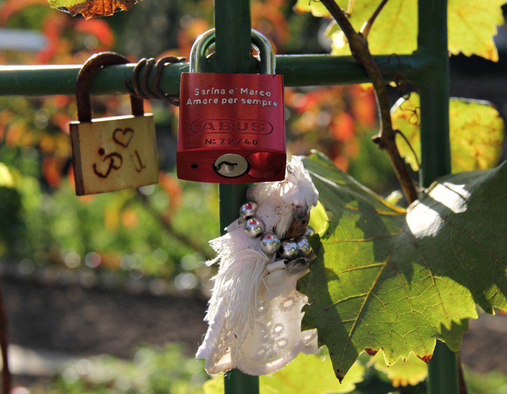 Zwei Liebesschlösser am Mehrgenerationengarten. Eines ist golden und graviert mit einem Herzen und den Buchstaben "P" und "U". Das andere ist Rot und trägt die Gravur "Sarina e Marco. Amore per sempre".