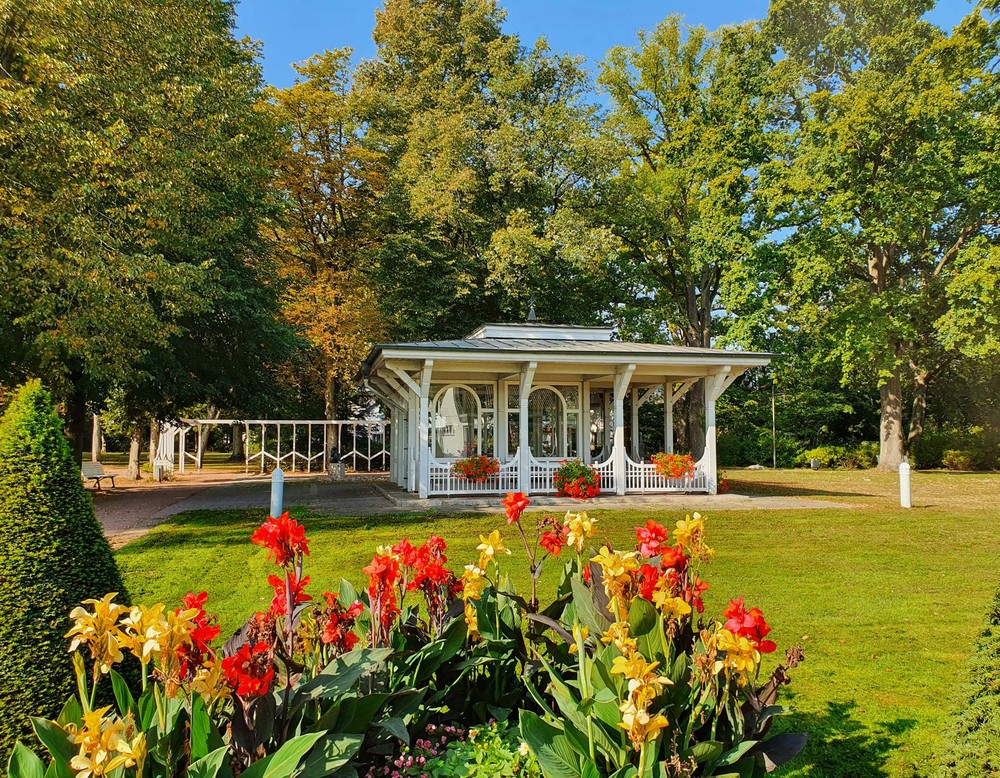 Parkhäuschen im Kurpark Bad Waldliesborn. Umgeben ist es von Bäumen und einer Wiese. Im Vordergrund sieht man Blumen.