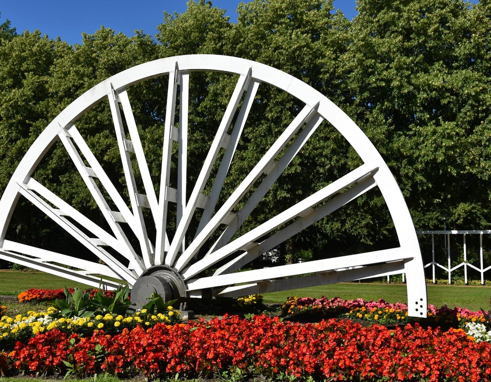 Kohlerad im Kurpark Bad Waldliesborn. Es ist von zahlreichen verschiedenfarbigen Blumen umgeben. Im Hintergrund stehen Bäumen.