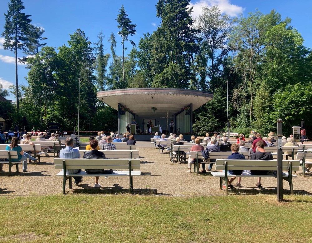 Frontale Ansicht der Konzertmuschel im Kurpark Bad Waldliesborn. Im Moment der Aufnahme fand ein Gottesdienst statt. Vor der Muschel stehen mehrere Bänke, auf denen Menschen sitzen.