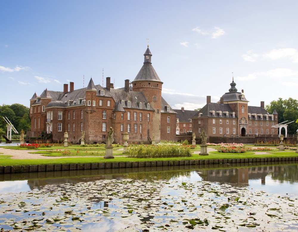 Blick über den Barockgarten des Parks auf die Westfassade der Wasserburg Anholt. Im Barockgarten stehen mehrere barocke Skulpturen. In den Beeten wachsen Rosensträucher.