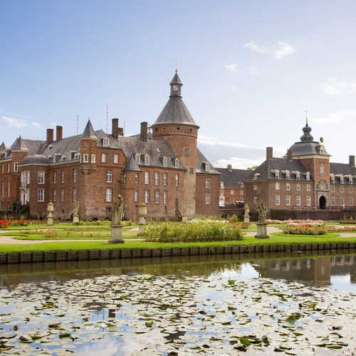 Blick über den Barockgarten des Parks auf die Westfassade der Wasserburg Anholt. Im Barockgarten stehen mehrere barocke Skulpturen. In den Beeten wachsen Rosensträucher.