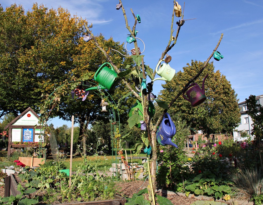 Ein Baum im Mehrgenerationengarten ist mit verschiedenen Gießkannen geschmückt. Umgeben ist der Baum von mehreren Beeten.