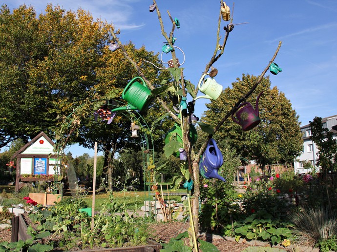 Ein Baum im Mehrgenerationengarten ist mit verschiedenen Gießkannen geschmückt. Umgeben ist der Baum von mehreren Beeten.