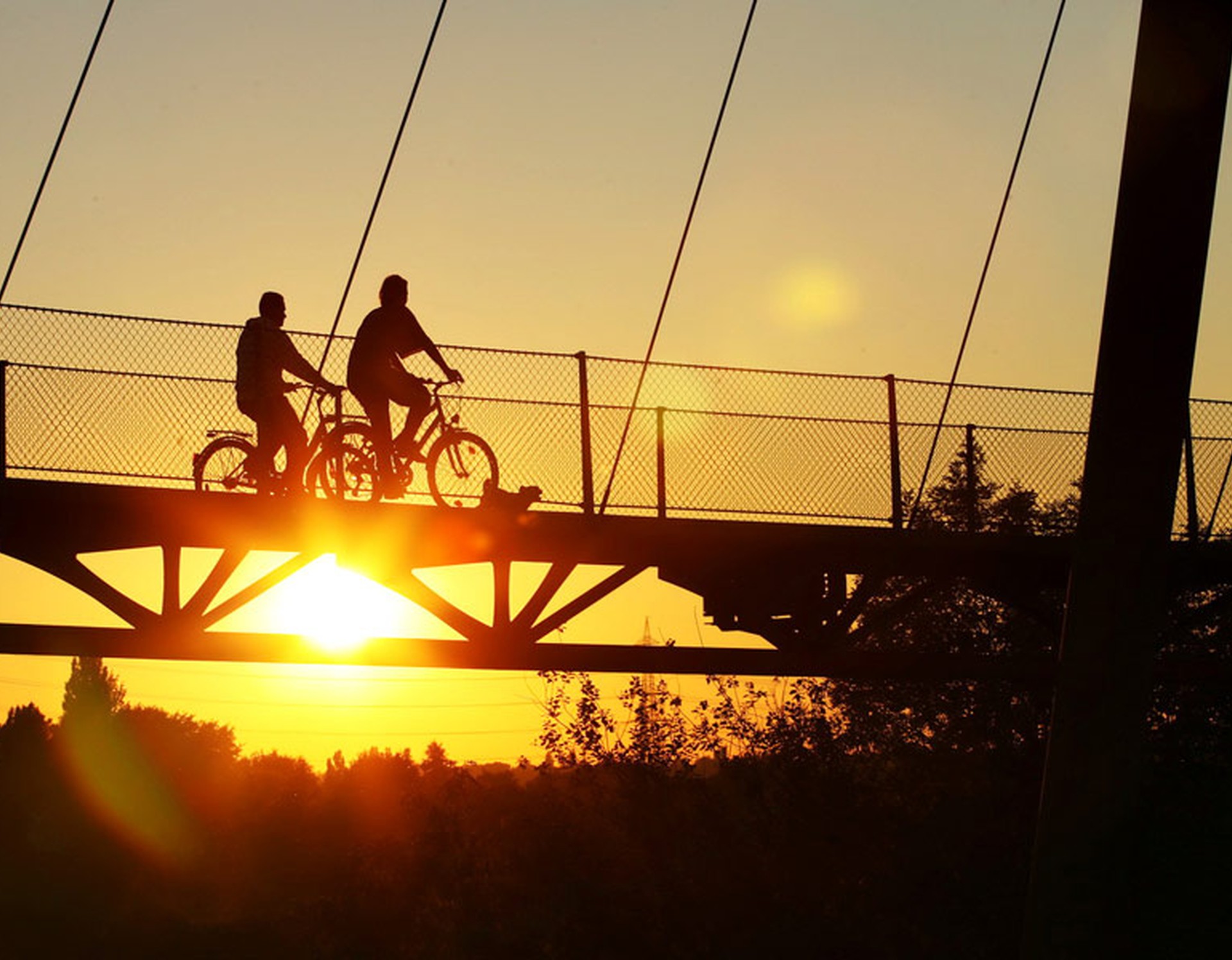 Brücke im Bochumer Westpark beim Sonnenuntergang. Diese wird von zwei Fahrradfahrern überquert.
