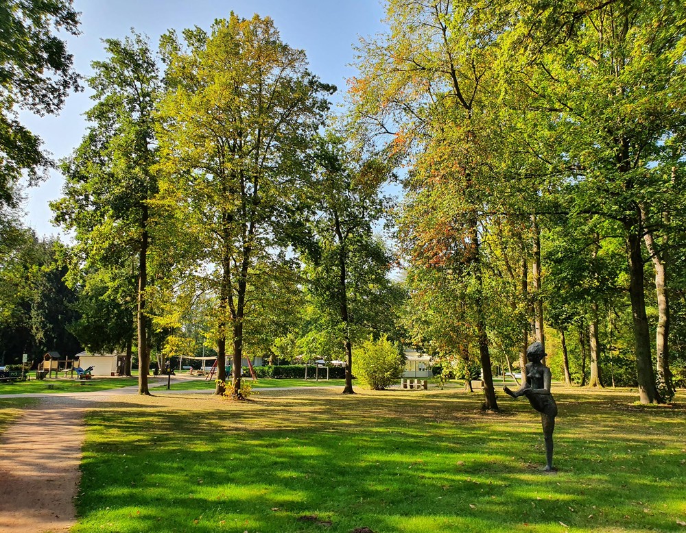 Blick in den Kurpark Bad Liesborn. Rechts sieht man eine der Bronzeskulpturen im Park, die auf einer Wiese steht. Rundherum stehen Bäume. Im Hintergrund sieht man die beiden Spielplätze.