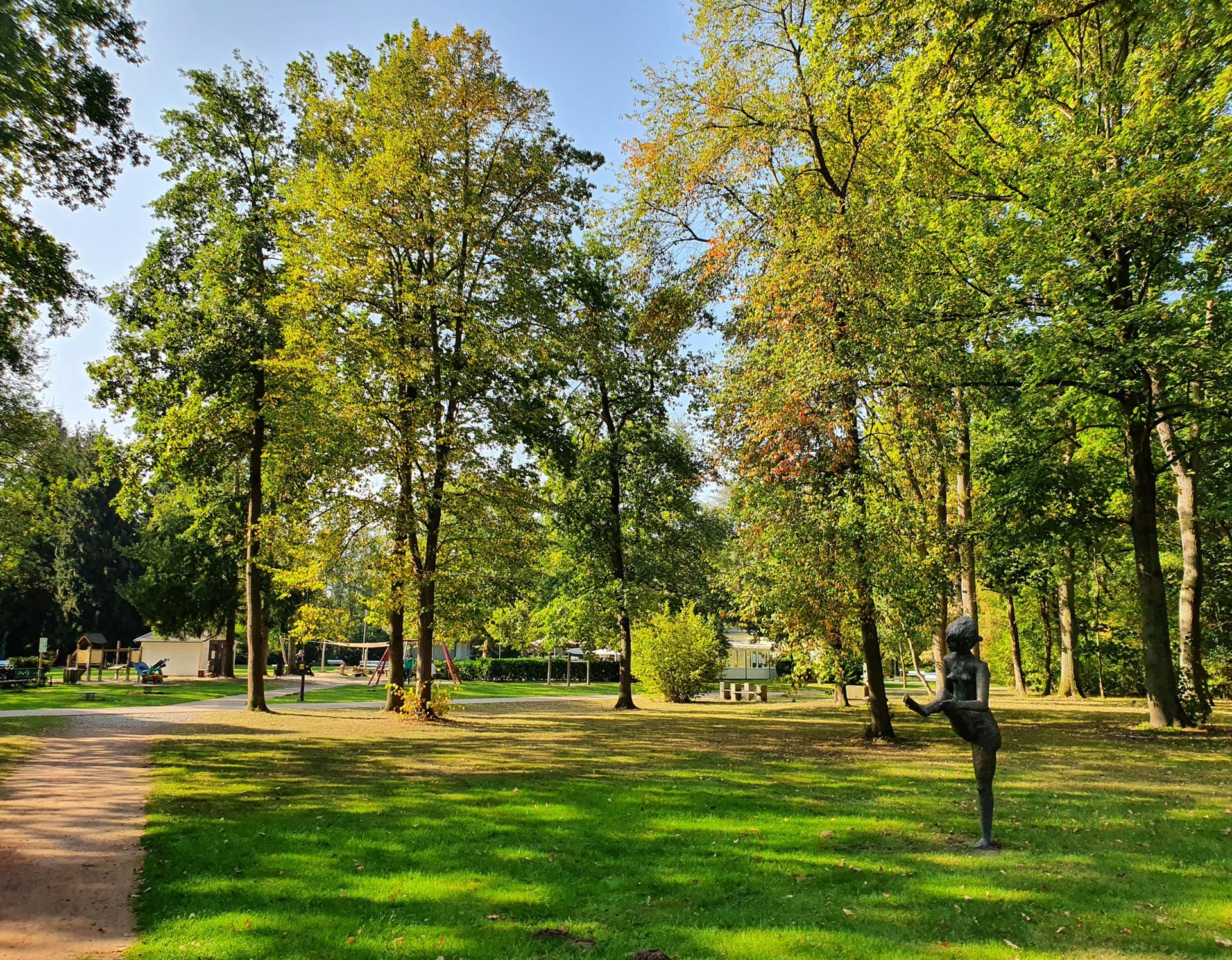 Blick in den Kurpark Bad Liesborn. Rechts sieht man eine der Bronzeskulpturen im Park, die auf einer Wiese steht. Rundherum stehen Bäume.