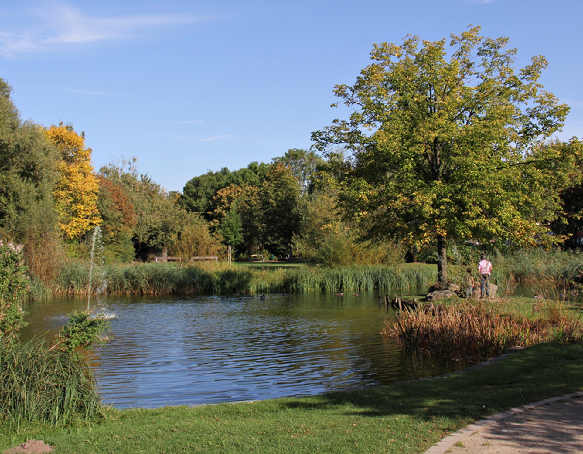 Ein Teich am Mehrgenerationengarten Lippstadt. Links befindet sich eine Fontaine. Auf einer kleinen Halbinsel steht ein Erwachsener mit einem Kind vor einem Baum.