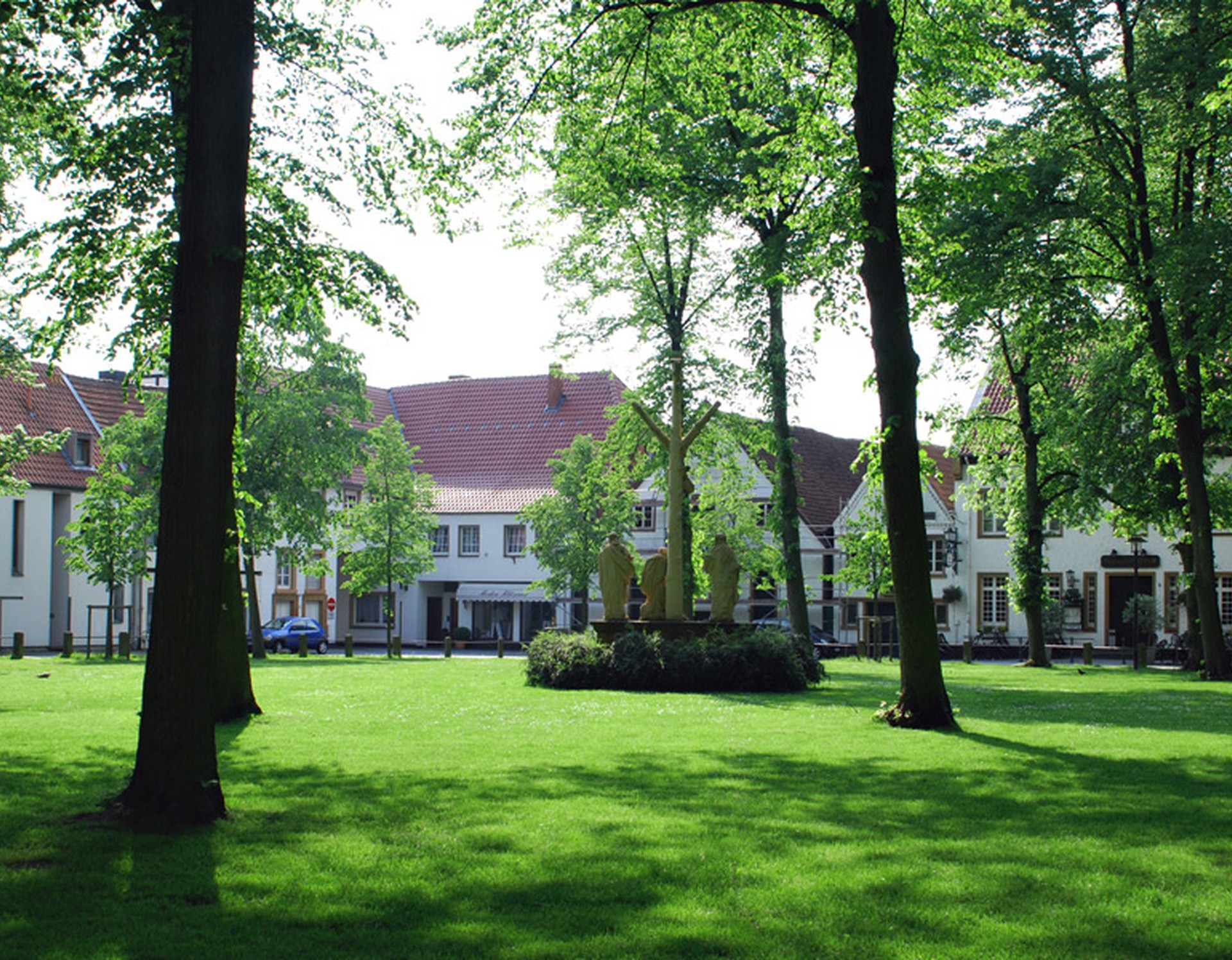 Der Kirchplatz am Kloster Herzebrock. In der Mitte befindet sich eine steinerne Kreuzigungsgruppe. Auf der Wiese des Platzes stehen mehrere Bäume.