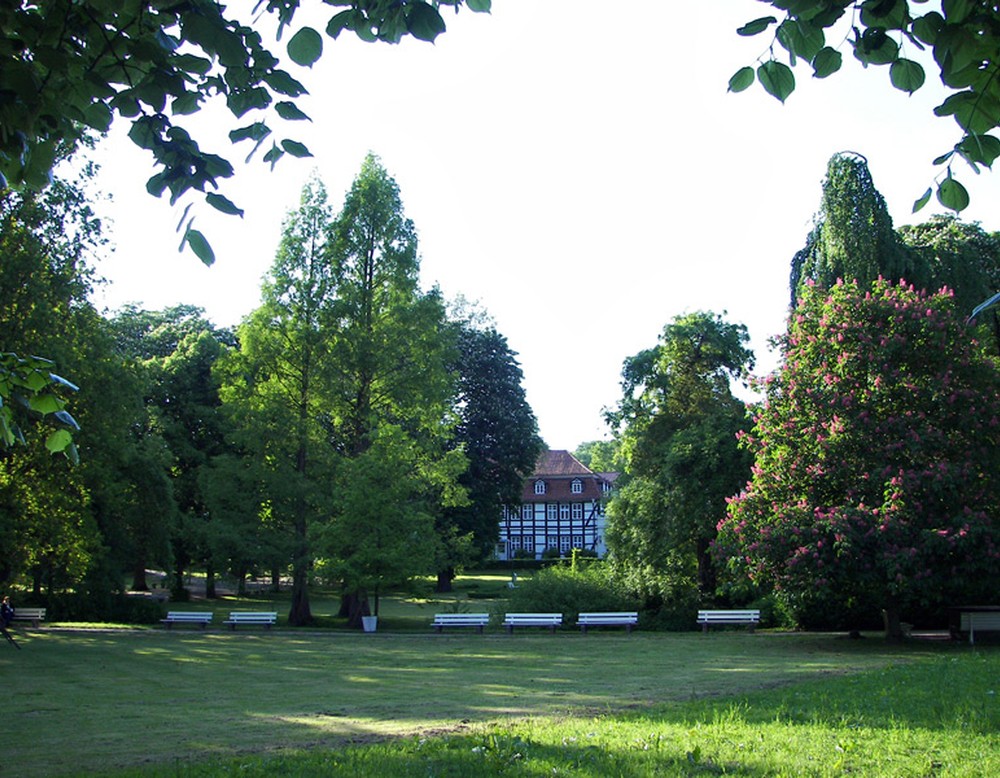 Eine Wiese im Kurpark Horn-Bad Meinberg. An dieser führt ein Weg mit mehreren Sitzbänken entlang. Im Hintergrund befindet sich ein Fachwerkhaus.