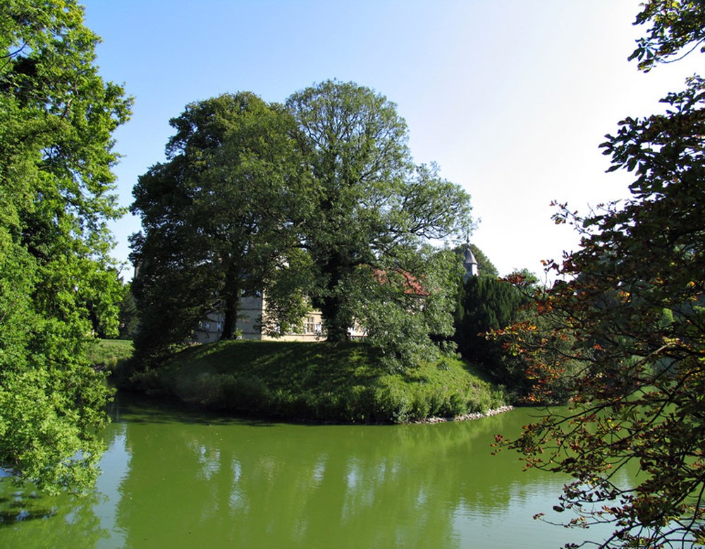 Einer der Wassergräben, die das Schloss Westerwinkel umgeben. Hinter dem Baum im Hintergrund ist das Hauptschloss zu sehen.